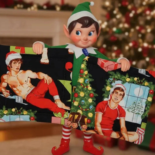Elf on the Shelf holding a Christmas-themed sweater with graphic designs in a festive room.